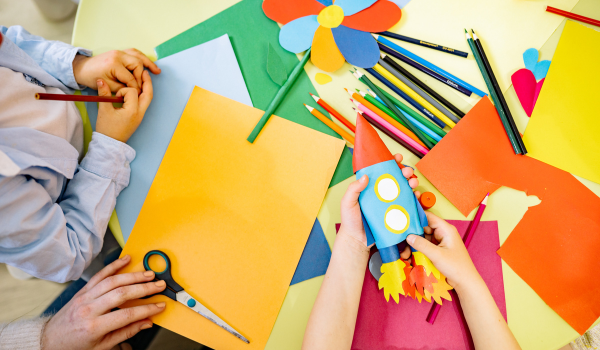 Children doing paper crafts with colored paper and pencils, with a rocket craft on the table