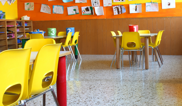 A bright classroom with yellow chairs and tables, with children’s drawings displayed on the wall