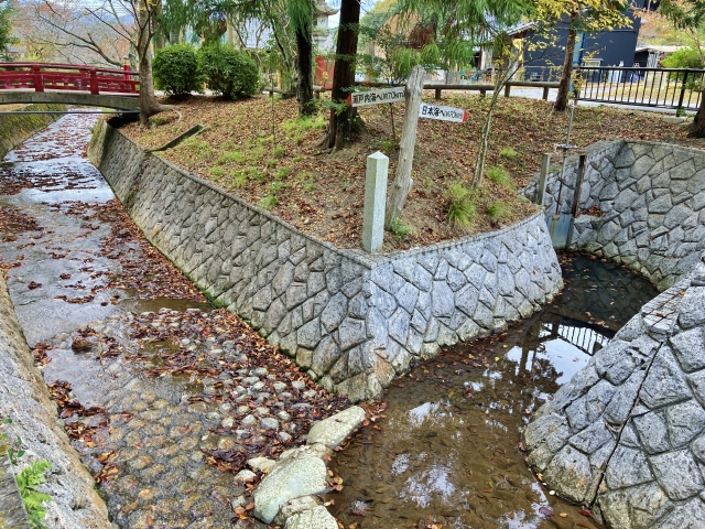 A stone-lined water channel and a red bridge at Mizuwakare Park with fallen leaves