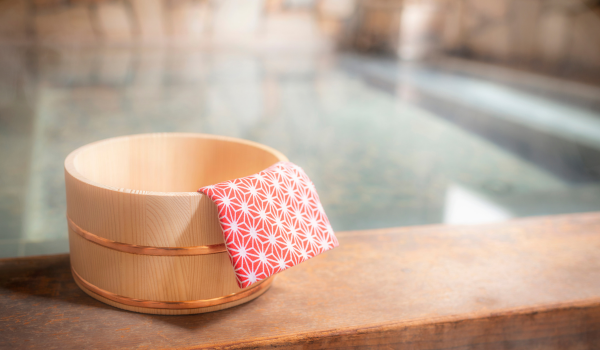 A wooden bath bucket and a red towel placed by a hot spring bath, conveying a relaxing onsen atmosphere